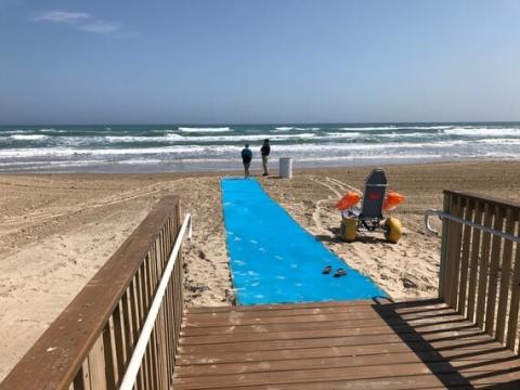 Picture of a Texas beach featuring a mobility mat and beach wheelchair with two people standing at the water's edge.